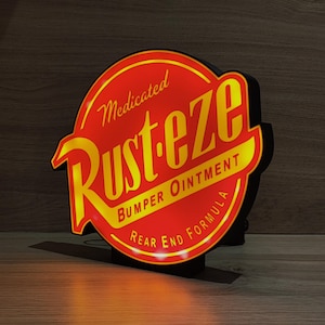Può includere: Un'insegna luminosa rossa e gialla con la scritta "Medicated Rusteze Bumper Ointment Rear End Formula". L'insegna ha un bordo nero ed è illuminata dall'interno. Forma circolare.