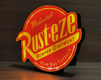 Op de afbeelding: Een verlicht bord met een rood en geel ontwerp. Het bord leest "Medicated Rusteze Bumper Ointment Rear End Formula" in gele tekst. Het bord heeft een zwarte rand en is van binnenuit verlicht.