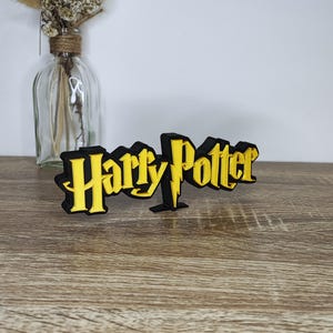 Peut inclure: Logo noir et jaune "Harry Potter" avec le texte "Harry Potter" dans une police stylisée. Le logo est sur une surface en bois. Un vase en verre transparent avec des fleurs séchées est en arrière-plan.