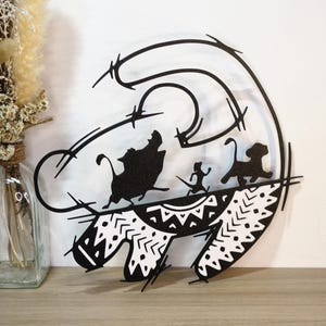 May include: A black and white decorative wall art piece featuring a stylised silhouette of a lion head outline containing smaller silhouettes of a warthog, a meerkat, and a lion cub walking, with a patterned base featuring geometric shapes.