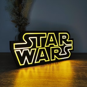 Könnte beinhalten: Ein schwarzes Star Wars-Schild mit gelber Neonbeleuchtung. Das Schild steht auf einer Holzoberfläche, und das Licht spiegelt sich auf der Oberfläche. Das Star Wars-Logo ist deutlich sichtbar.