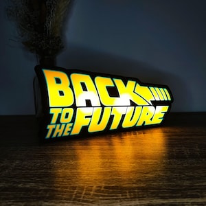 Può includere: Insegna luminosa con la scritta "BACK TO THE FUTURE" in giallo e verde acqua. L'insegna rettangolare ha un bordo nero ed emette una luce calda, che si riflette sulla superficie sottostante. Un oggetto nostalgico, che fa riferimento all'iconico film.