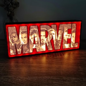 Op de afbeelding: Een rechthoekig, verlicht bord met het woord "MARVEL" in witte letters op een rode achtergrond. De letters zijn gevuld met afbeeldingen van Marvel-personages. Het bord straalt een warm licht uit, wat een opvallend visueel effect creëert.