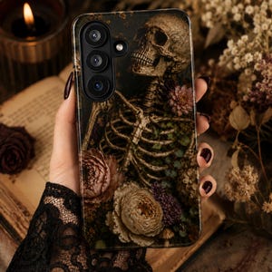 May include: A rectangular smartphone case featuring a dark, detailed graphic of a human skeleton intertwined with various muted-toned flowers, including large beige roses and small purple blossoms, set against a dark, moody background.