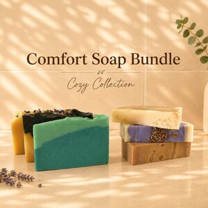 May include: A collection of rectangular soap bars in shades of teal, yellow, black, white, purple, and brown, with some bars topped with dried lavender. The text Comfort Soap Bundle Cozy Collection is displayed above the soaps.
