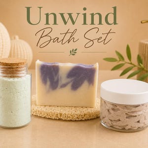 Puede incluir: Un juego de baño con el texto "Unwind Bath Set". El juego incluye un tarro de sales de baño, una pastilla de jabón con remolinos morados y un tarro de exfoliante corporal. Un dispensador de jabón decorativo y objetos decorativos están en el fondo.