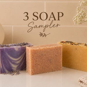 Könnte beinhalten: Ein Seifen-Sampler mit drei handgemachten Seifenstücken. Die Seifen sind in Lila-, Braun- und Gelbtönen gehalten und weisen sichtbare Texturen und botanische Elemente auf. Der Text "3 SOAP Sampler" ist über den Seifen zu sehen.