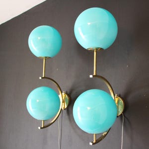May include: Four-light wall sconces with turquoise globe shades. The sconces have a brass finish and a curved design. The globes are a bright, opaque turquoise color. The sconces are mounted on a dark gray wall.