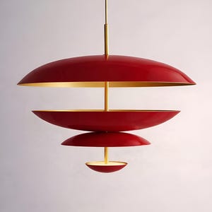 May include: A modern, tiered pendant light fixture. The light features a series of red, saucer-shaped shades with gold-colored interiors. A central gold-colored rod suspends the shades. The fixture is designed to be hung from the ceiling.