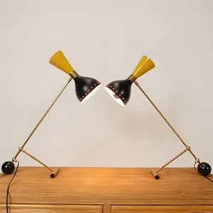 Op de afbeelding: Twee bureaulampen in mid-century modern stijl. Elke lamp heeft een gele conische kap, een zwarte behuizing en een messingkleurige arm en voet. De lampen staan op een houten oppervlak, met een zwarte bol aan de voet van elke lamp.