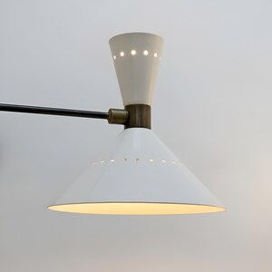 May include: A white and cream-colored mid-century modern wall sconce. The sconce features a conical shade with small perforations, a cream-colored top, and a black arm. The lamp has a brass-colored joint and is designed to be mounted on a wall.