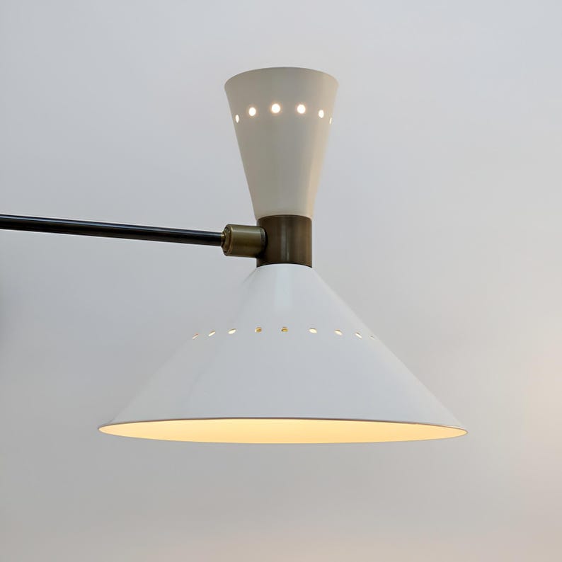 May include: A white and cream-colored mid-century modern wall sconce. The sconce features a conical shade with small perforations, a cream-colored top, and a black arm. The lamp has a brass-colored joint and is designed to be mounted on a wall.