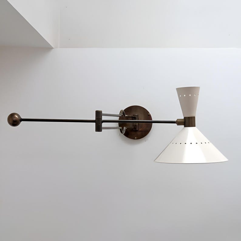 May include: A wall-mounted light fixture with a white conical shade and a cream-colored upper section. The fixture has a dark metal arm and a circular base. The shade has small, evenly spaced holes.