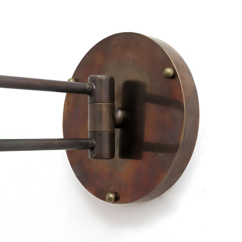 May include: A wall-mounted, bronze-colored light fixture with a circular backplate and two adjustable arms. The fixture has a vintage aesthetic, with visible screws and a warm, metallic finish. The arms extend from a central hinge.
