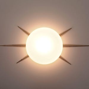 Op de afbeelding: Een moderne plafondlamp met een grote, gloeiende witte bol in het midden, omringd door acht goudkleurige, puntige steruitbarstingselementen. De armatuur is op een neutraal gekleurd plafond gemonteerd.