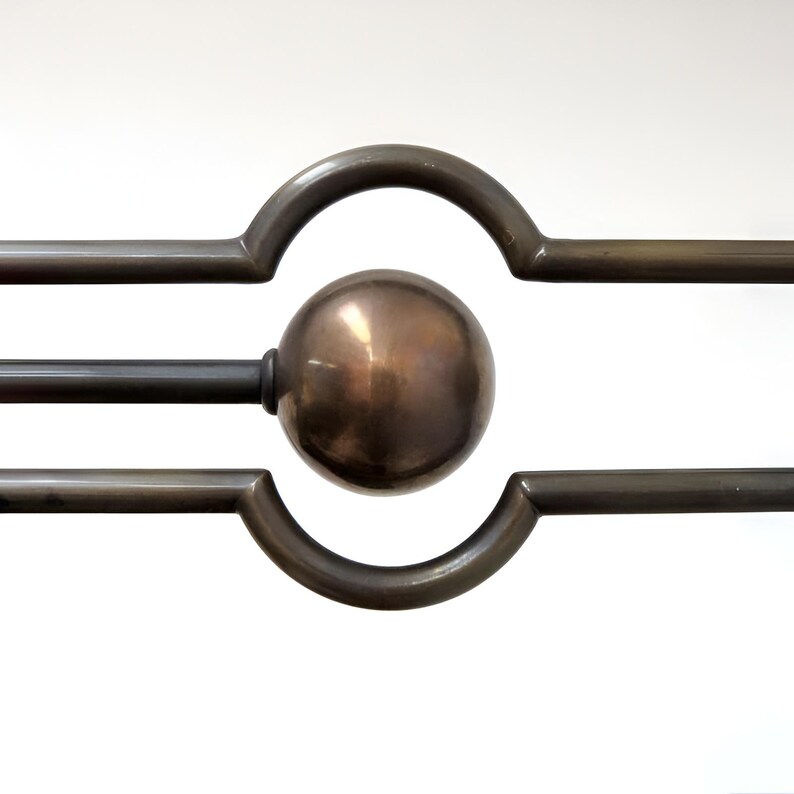 May include: Close-up of a bronze-colored metal curtain rod with a spherical finial. The rod features a decorative design with curved and straight lines, creating a geometric pattern. The finish has a warm, metallic sheen.