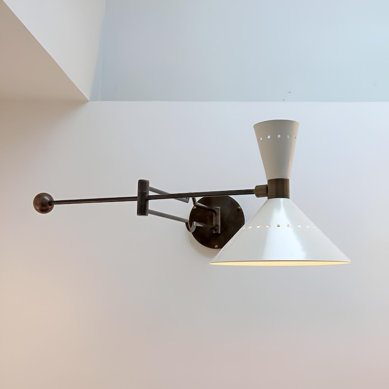 May include: A white and gray wall-mounted lamp with a cone-shaped shade and a pivoting arm. The lamp has a dark bronze finish on the arm and mounting plate. The shade has small perforations near the top.
