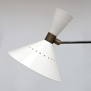 May include: A white, cone-shaped lamp with a brass-colored joint and a black arm. The lamp has small holes around the edge of the shade. The lamp is angled, with the light facing upwards. The lamp is a modern design.
