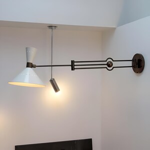 May include: A modern wall-mounted lamp with a white conical shade and a smaller cylindrical light. The lamp features a dark metal arm and a circular wall mount. The design is minimalist and stylish, suitable for interior lighting.