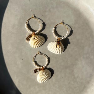 Op de afbeelding: Drie paar gouden oorringen. Elke oorring heeft een witte schelpenhanger, een rij kleine witte kralen en een gouden initiaalbedel. De oorbellen worden gepresenteerd op een lichtgrijze ondergrond.