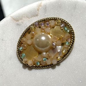 Op de afbeelding: Ovale broche met een goudkleurige rand. Het midden is versierd met een grote, ronde parel, omringd door kleine, iriserende kralen in roze, geel en groen. De broche heeft een vintage uitstraling.