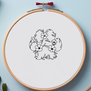 May include: A wooden embroidery hoop with a white fabric featuring a black floral paw print design. The hoop is surrounded by green and yellow leaves and other craft supplies on a light blue surface.