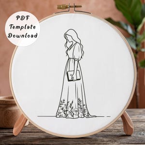 Puede incluir: Un bastidor de bordado circular de madera con tela blanca y una ilustración de líneas negras de una persona con un vestido largo sosteniendo un libro. El dobladillo del vestido tiene un patrón floral. El texto PDF Template Download es visible.