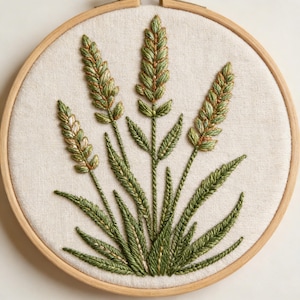 Op de afbeelding: Een geborduurd kunstwerk met groene en gouden tarwearen op een crèmekleurige stof, geplaatst in een houten borduurring. De tarwearen zijn ingewikkeld gedetailleerd met verschillende tinten groen en goud garen, waardoor een gestructureerd, botanisch ontwerp ontstaat.