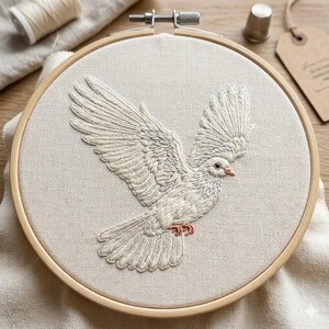 以下が含まれることがあります： ベージュ色の布に施された、白い鳩の刺繍。木製の枠に張られており、羽を広げた姿が細かく表現されています。鳩のくちばしと足はソフトコーラル色です。背景には、糸や指ぬきなどの裁縫道具が写っています。
