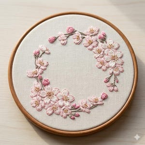 May include: A handcrafted embroidery hoop featuring a wreath of pink cherry blossoms. The flowers are intricately stitched on a cream-colored fabric, set within a round wooden frame. The design includes delicate buds and open blossoms.