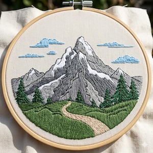 May include: Embroidered mountain landscape in a wooden hoop. The design features a gray mountain range with snow-capped peaks, green trees, and a winding path. Blue clouds dot the sky, and the grass is a vibrant green.