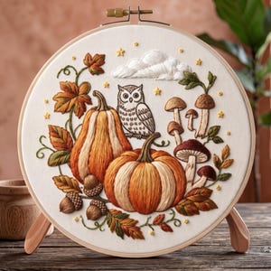 Può includere: Un telaio da ricamo circolare contenente un pannello in tessuto bianco con un dettagliato disegno ricamato raffigurante due zucche arancioni, un gufo grigio, funghi marroni, ghiande e foglie autunnali, accentuato da piccole stelle gialle.