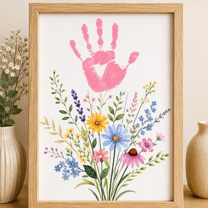 以下が含まれることがあります： ピンク色の手形が描かれ、その下に青、黄、ピンクの花々と緑の茎や葉が色鮮やかに描かれた野花のイラストが配置された、明るい木製フレーム入りのプリント作品。