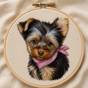 May include: A circular wooden embroidery hoop containing a detailed thread-stitched portrait of a small dog with black, tan, and grey fur. The dog is wearing a textured pink scarf around its neck, set against a plain, light-colored fabric background.