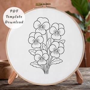 May include: A circular wooden embroidery hoop containing white fabric with a black line art illustration of a pansy flower bouquet. The text "PDF Template Download" is printed in black on the upper left side of the white fabric surface.