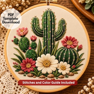 May include: An embroidery hoop containing a fabric panel with a stitched cactus and flower design. The artwork features green cacti with white spines, pink and white flowers, and small brown rocks. Text reads: PDF Template Download and Stitches and Color Guide Included.