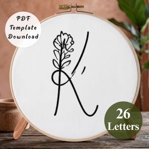 May include: A circular wooden embroidery hoop containing white fabric with a black line art illustration of the letter K integrated with a floral stem. A circular white label reads PDF Template Download and a green circle reads 26 Letters.