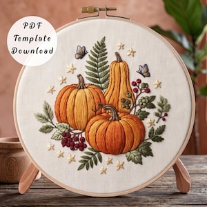 Può includere: Un telaio da ricamo contenente un pannello in tessuto con un disegno ricamato di due zucche arancioni, una zucca allungata, foglie di felce verdi, bacche rosse e piccole farfalle blu. Il testo "PDF Template Download" è stampato nell'angolo in alto a sinistra.