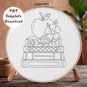 May include: A round wooden embroidery hoop containing white fabric with a black line art design of a stack of three books topped with an apple and a flower. The text PDF Template Download is visible in the upper left corner of the image.