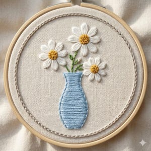 May include: Hand-stitched embroidery art featuring a light blue vase holding three white daisies with yellow centers. The embroidery is set in a wooden hoop, with a detailed border. The background is a neutral fabric.