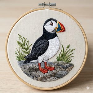 May include: Embroidered artwork of a puffin bird. The bird has a black and white body, a large colorful beak, and red feet. It is perched on a gray rock with green grass, all within a wooden embroidery hoop.