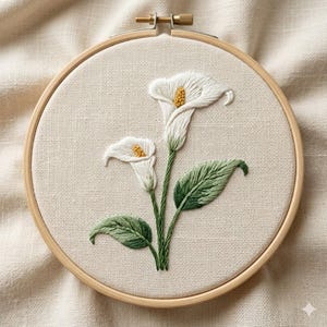 May include: A circular wooden embroidery hoop containing a piece of off-white fabric. The fabric features a hand-stitched design of two white calla lilies with yellow centers, accompanied by green stems and textured green leaves.