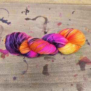 Peut inclure: Une écheveau de fil coloré aux tons de violet, rose, orange et jaune. Le fil est disposé en forme torsadée et enroulée, avec les couleurs qui se fondent. Ce fil est probablement utilisé pour des projets de tricot ou de crochet.