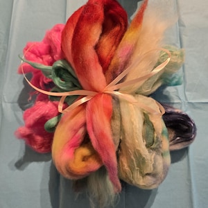 Könnte beinhalten: Ein Bündel farbenfroher Wollrovinge, mit Farbtönen von Rosa, Orange, Gelb und Blau, gebunden mit einem hellrosa Band. Die Wolle ist weich und flauschig, ideal für Textilkunstprojekte wie Spinnen oder Filzen.
