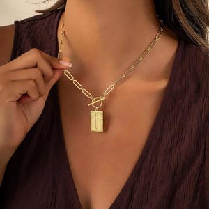 May include: Gold-colored necklace featuring a rectangular pendant with a cross design. The pendant is attached to a chain with a toggle clasp. The necklace is a religious-themed accessory.