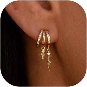 May include: Gold-coloured triple hoop earrings. The top hoop is encrusted with small, clear crystals. Two additional hoops dangle below, each with a decorative charm. One charm is a small, pointed shape, and the other has a chain of crystals.