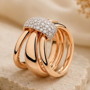Könnte beinhalten: Ein mehrreihiger Ring aus poliertem Roségold mit einem breiten, geschwungenen Mittelsteg, der mit zahlreichen kleinen, klaren, rund geschliffenen weißen Diamanten besetzt ist und über die glatten, abgerundeten Metallbänder verläuft.