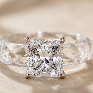 Puede incluir: Un anillo transparente y facetado con una gran piedra central de corte cuadrado sujeta por cuatro garras metálicas. La banda está hecha de un material transparente y texturizado con un patrón geométrico similar a un diamante.