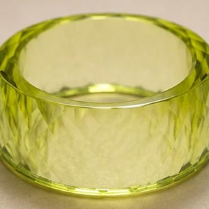 May include: A wide, translucent lime green bangle bracelet featuring a faceted, diamond-cut exterior surface. The circular accessory has a smooth interior band and a reflective, geometric pattern across the outer circumference of the piece.