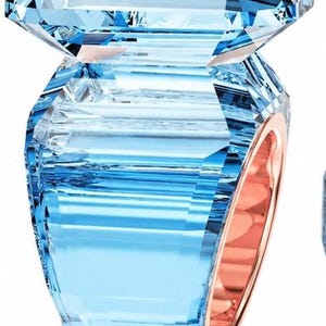 Könnte beinhalten: Ein Ring mit einem großen, facettierten, hellblauen Kristallstein auf einem breiten, geometrischen, hellblauen Kristallband. Die Innenseite des Bandes ist mit einer glatten, polierten Roségold-Metalloberfläche versehen, die durch den Kristall sichtbar ist.